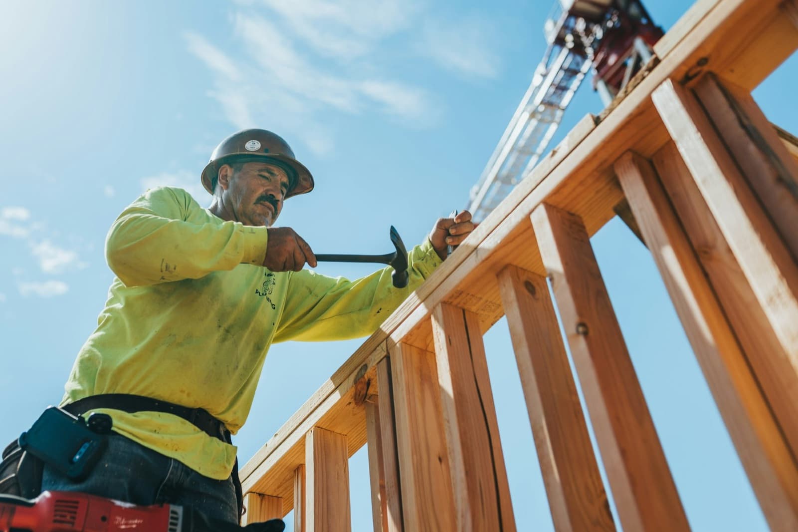 A carpenter in a hard hat hammering the top plate of a new stud-framed wall against a blue sky